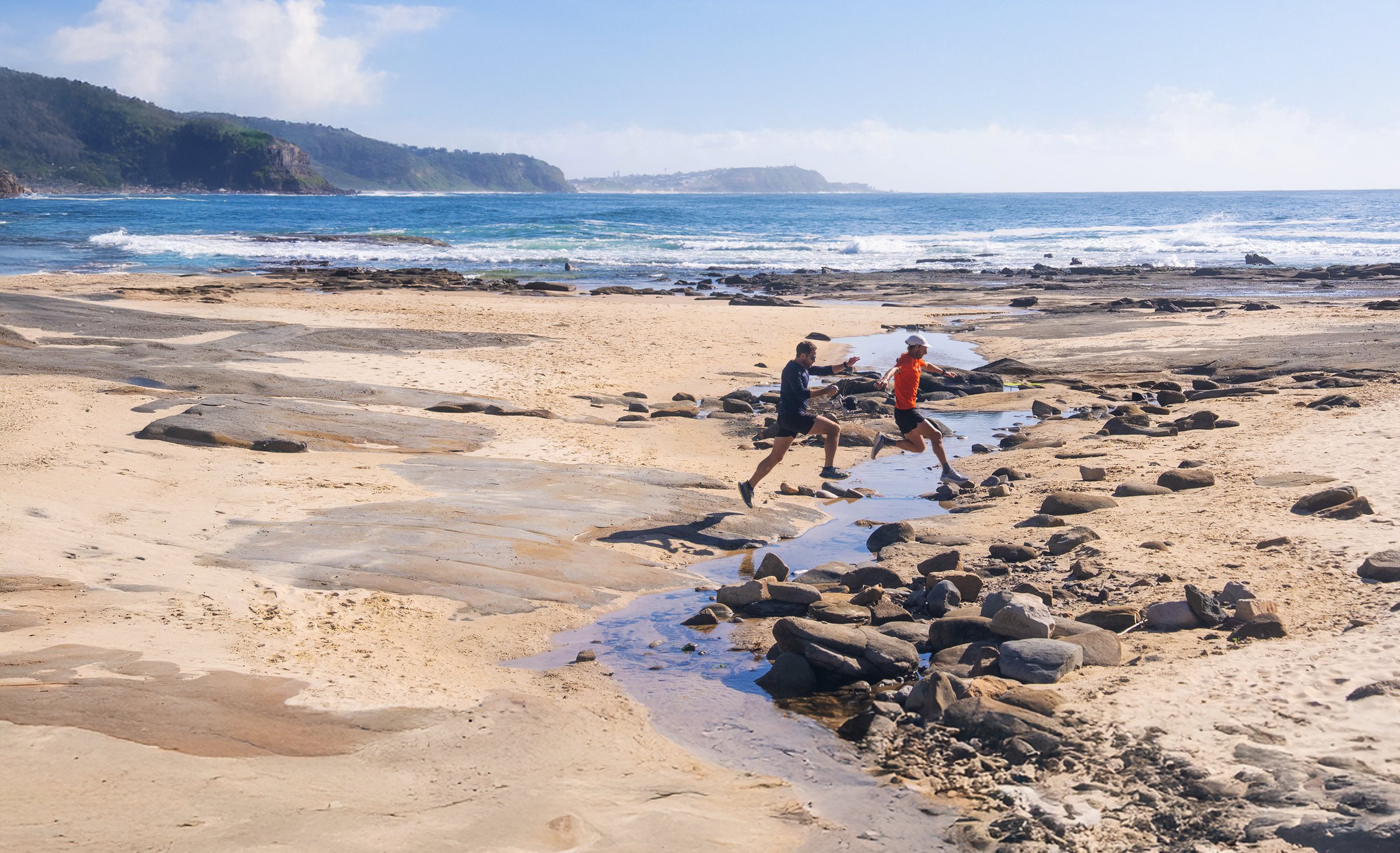 Chris and Vlad jumping a stream on a Newcastle beach with cliffs in the distance